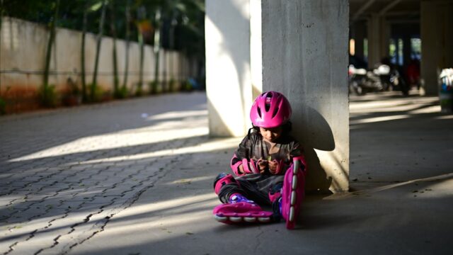 Young girl in pink helmet with rollerblades sits by pillar