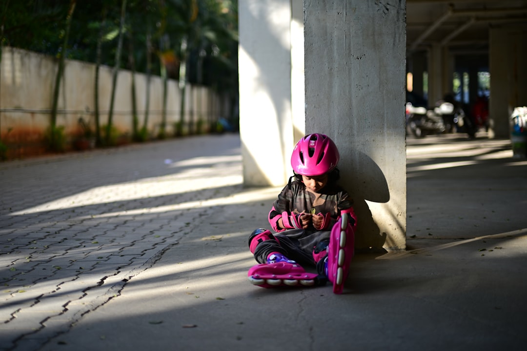 Young girl in pink helmet with rollerblades sits by pillar